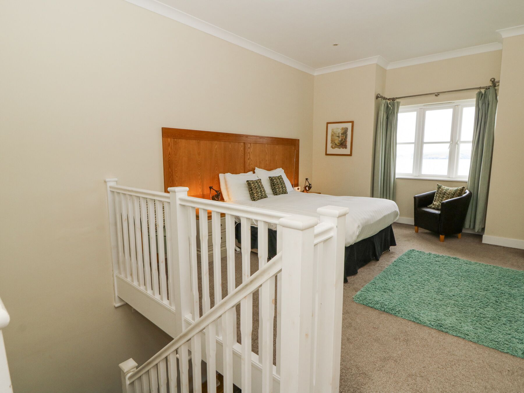 A bedroom with a wooden headboard, a bed with white bedding, a green area rug, a chair, and a window with curtains.