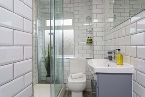 Et moderne badeværelse med hvide fliser, en glasbrusekabine, toilet og håndvask med toiletartikler.
