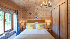 Ein Schlafzimmer einer Holzhütte mit Bett, Nachttischlampen, einer Chandelierleuchtre und einem Fenster mit Vorhängen.