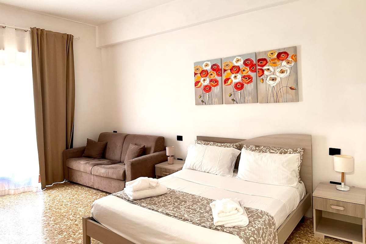A hotel room with a bed, a sofa, a nightstand, and a painting of flowers on the wall.