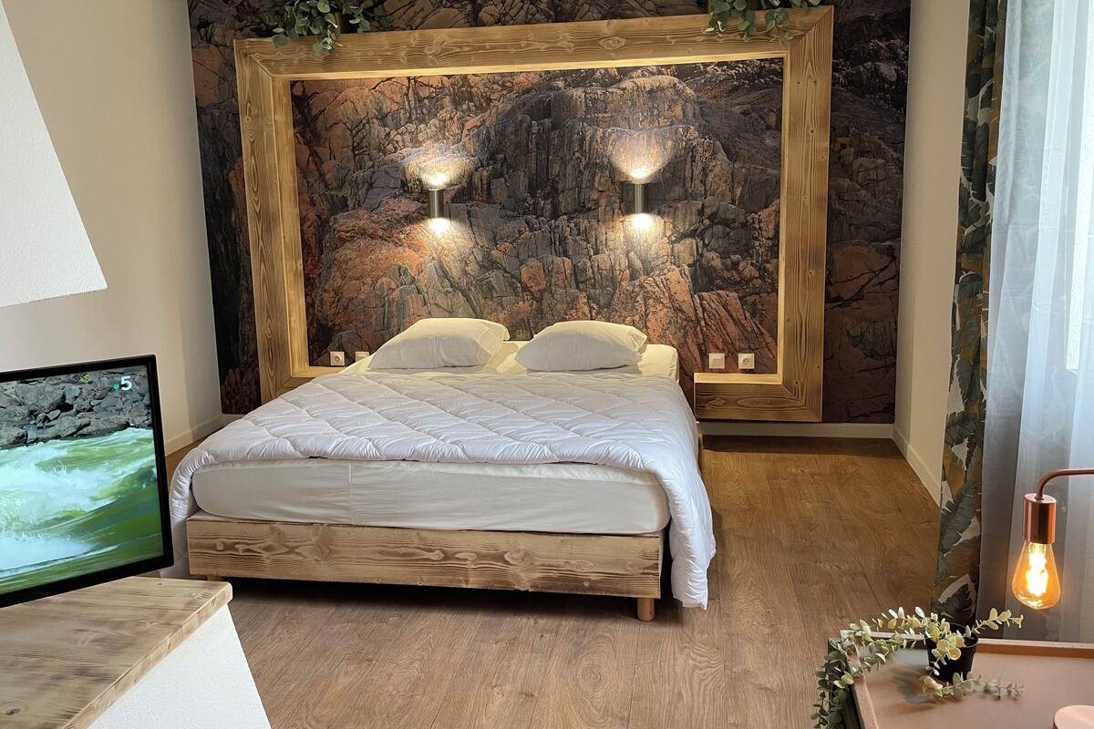 A bedroom with a bed, a wooden headboard, a wall mural, two wall-mounted lights, a bedside table, a TV, and a potted plant.