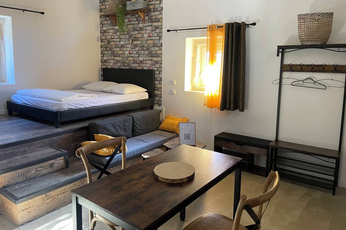 A compact room with a bed, a small sofa, a dining table, and a window with curtains.