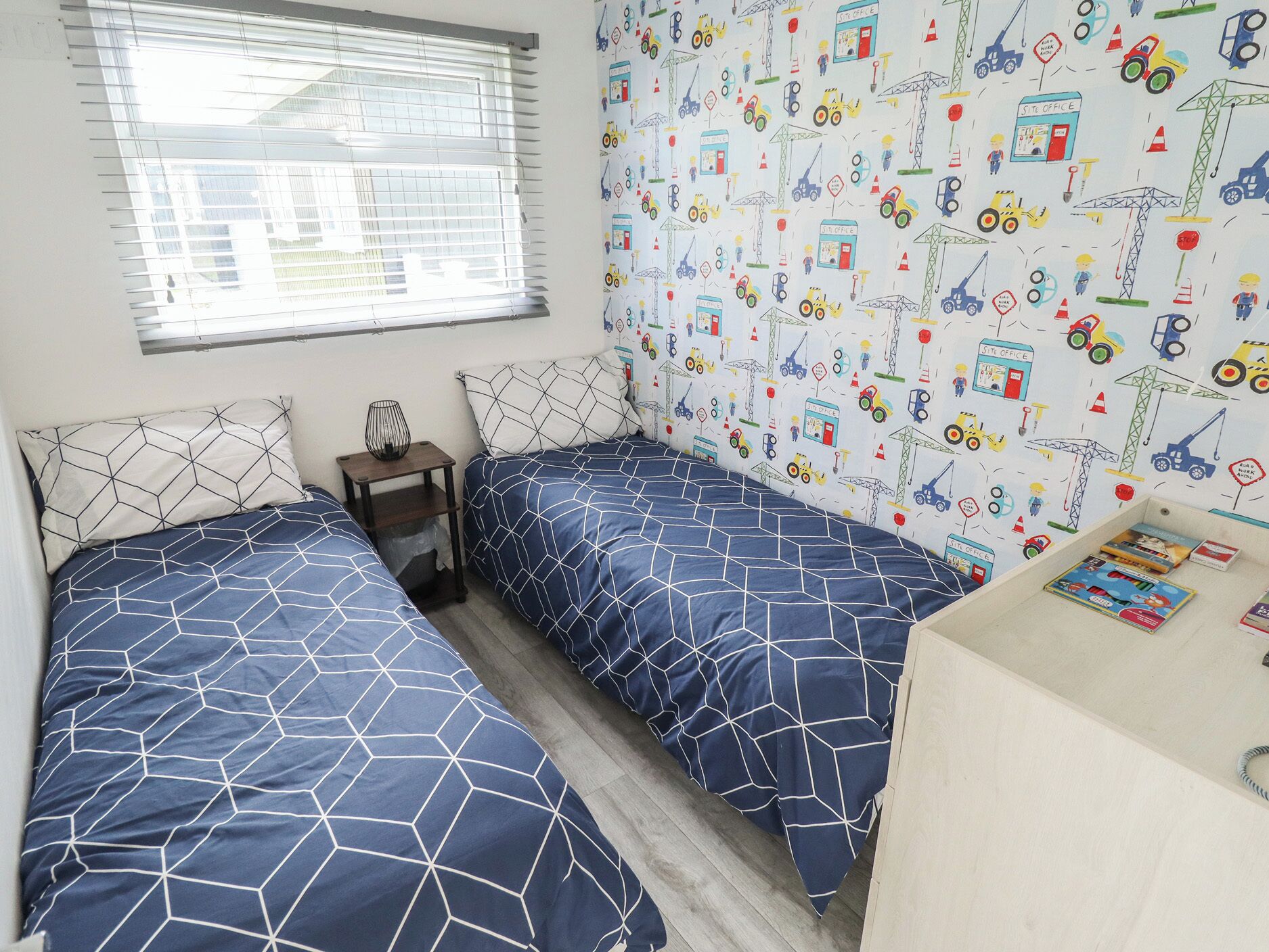 Un dormitorio con dos camas, una mesita de noche pequeña, una ventana con persianas y un papel tapiz con motivos de construcción.