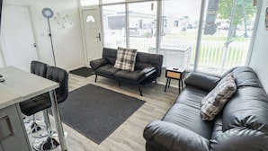 Un salon moderne avec un canapé, une table à manger et un tabouret de bar. On y trouve une grande fenêtre avec des stores, une lampe sur pied et une porte.
