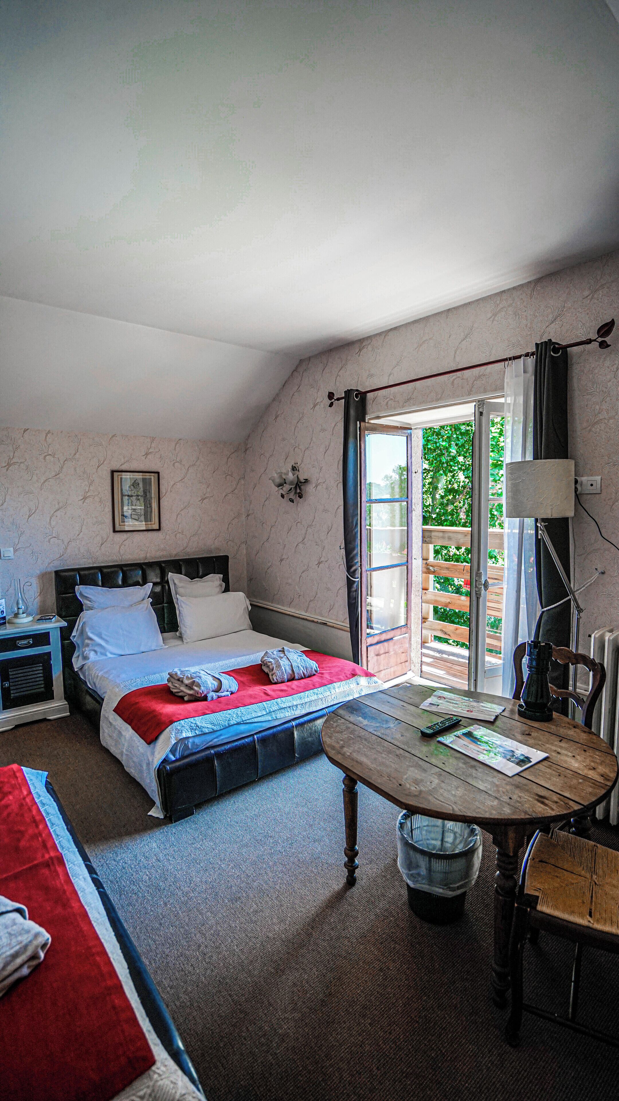 A bedroom with a bed, a table, a chair, a nightstand, and a window with a view of greenery.