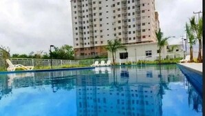A swimming pool with clear blue water, surrounded by a white fence, and a tall residential building in the background. - APARTMENT SEA BRISE ARAÇAGY (Araçagi São José de Ribamar)