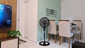 A modern living room with a flat-screen TV, a standing fan, a dining area with two chairs, and a wall with decorative art. - APARTMENT SEA BRISE ARAÇAGY (Araçagi São José de Ribamar)
