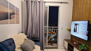 A living room with a sofa, a TV, and a sliding glass door leading to a balcony. - APARTMENT SEA BRISE ARAÇAGY (Araçagi São José de Ribamar)