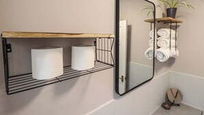 Un bagno con uno scaffale in legno con asciugamani, un porta carta igienica in metallo nero e uno specchio che riflette una pianta e degli asciugamani.