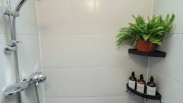 A shower with a handheld showerhead and a wall-mounted faucet, a potted plant on a black shelf, and a black shelf with bottles of shampoo and conditioner.