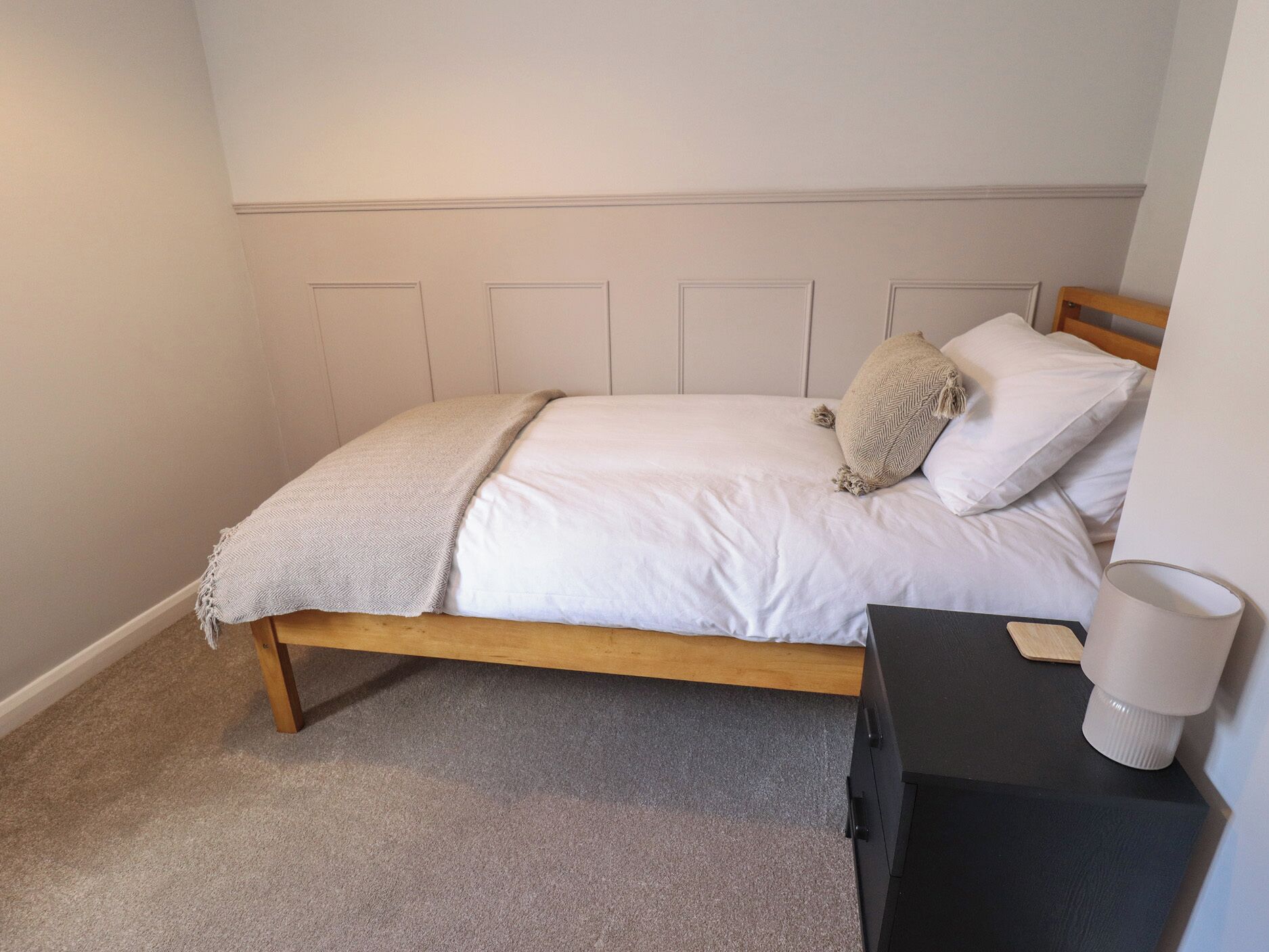 A bedroom with a wooden bed, white bedding, a beige throw blanket, and a small bedside table with a lamp.