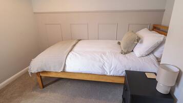 A bedroom with a wooden bed, white bedding, a beige throw blanket, and a small bedside table with a lamp.