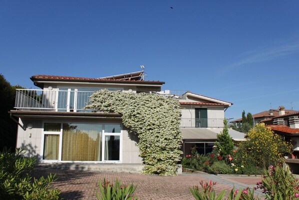 Una casa de dos pisos con un balcón, un ventanal grande y un jardín con diversas plantas y flores.