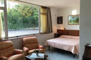Una habitación con una cama, dos sillones a rayas, una mesita con un florero, una ventana con cortinas y un cuadro en la pared.