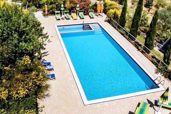 Una piscina rectangular con superficie de agua azul cristalina, rodeada por una solana con baldosas. Hay sillas de descanso y sombrillas a lo largo de la piscina.