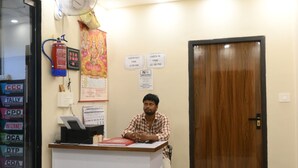 A reception area with a reception desk, a fire extinguisher, a wall-mounted fan, and a door with a sign indicating check-out times. - Shree Hari Niwas Paying Guest House (Varanasi)