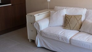 A white sofa with a beige pillow, a white bedside table with a lamp, and wooden cabinets. - Le Calypso - Magnificent sea-view terrace - 100m from Le Boucanet beach (Le Grau-du-Roi)