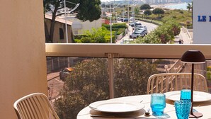 A balcony with a table set for two, overlooking a coastal road with parked cars and greenery. - Le Calypso - Magnificent sea-view terrace - 100m from Le Boucanet beach (Le Grau-du-Roi)