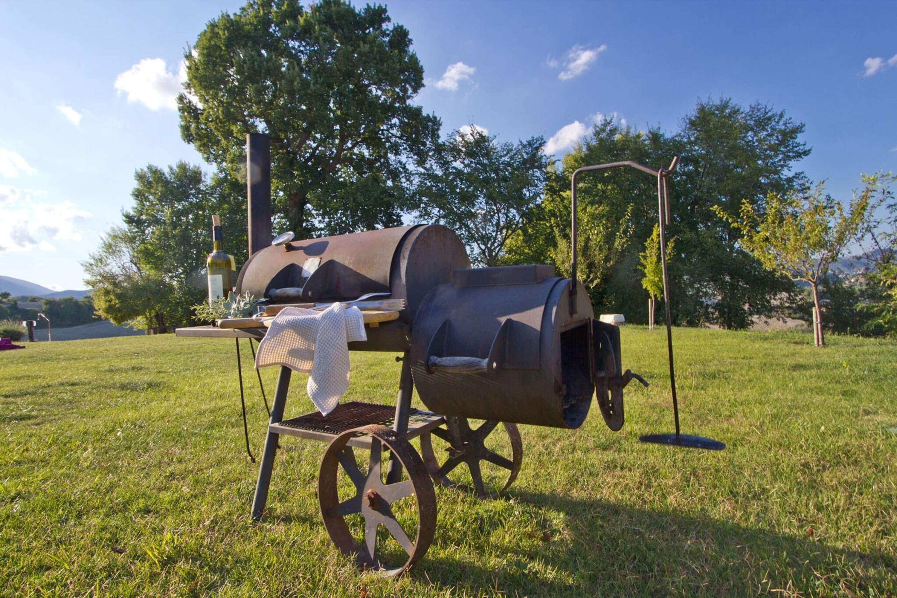 Una parrilla portátil con una funda de tela, sobre un campo cubierto de césped con árboles y un cielo despejado al fondo.