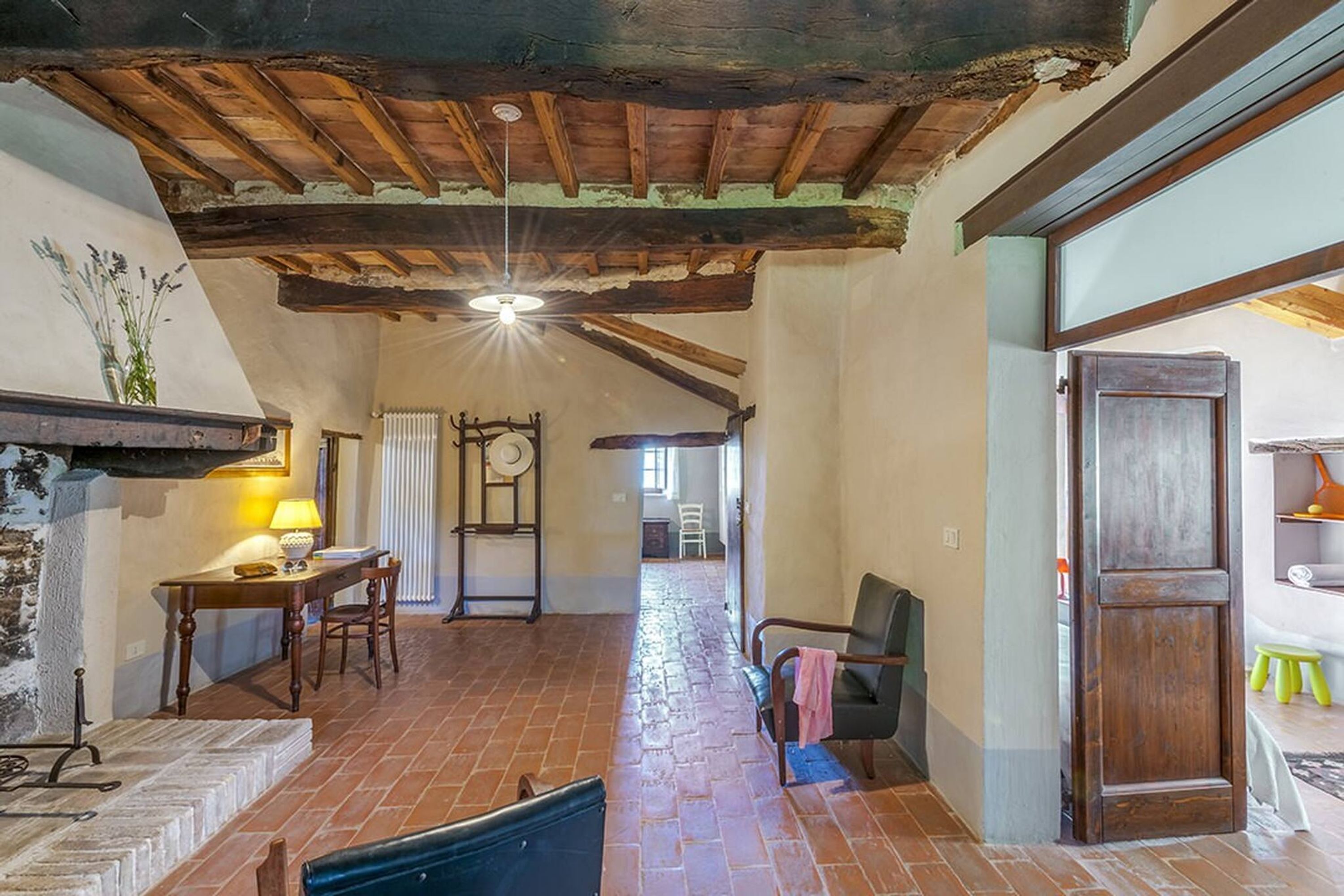 A rustic interior with exposed wooden beams, a brick floor, a fireplace, and a dining area with a table and chairs.