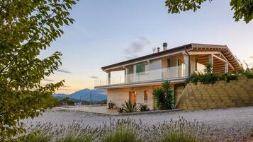 A modern two-story house with a balcony, a driveway, and a view of mountains in the background.