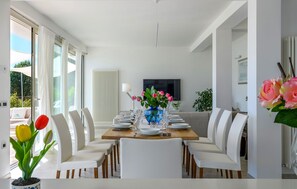 Une salle à manger moderne avec une table en bois dressée pour six personnes, des chaises blanches, un vase rempli de fleurs et un téléviseur en arrière-plan.