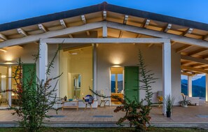 A covered outdoor area with a wooden ceiling, a seating area with chairs and a table, and a view of a mountain range.