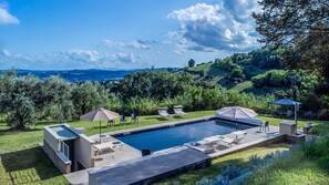Ein rechteckiger Swimmingpool mit Liegestühlen und Sonnenschirmen, umgeben von einer angelegten Gartenanlage und Blick auf ferne Hügel.