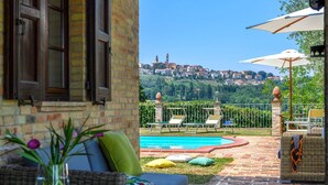 A patio with a pool, wicker furniture, and a view of a town in the background. - La Girolima 6 2 in Mondavio (Mondavio)