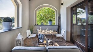 A balcony with a table set for two, white chairs, a potted plant, and a view of greenery. - Casale Degli Ulivi 8 2 (Acqualagna)