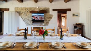 Une salle à manger avec un mur en pierre, des poutres apparentes, un téléviseur et une table dressée pour six personnes, agrémentée de bouteilles de vin et de verres.