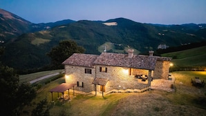 Une maison en pierre avec un toit de tuiles, entourée d’un paysage verdoyant et de collines en arrière-plan.