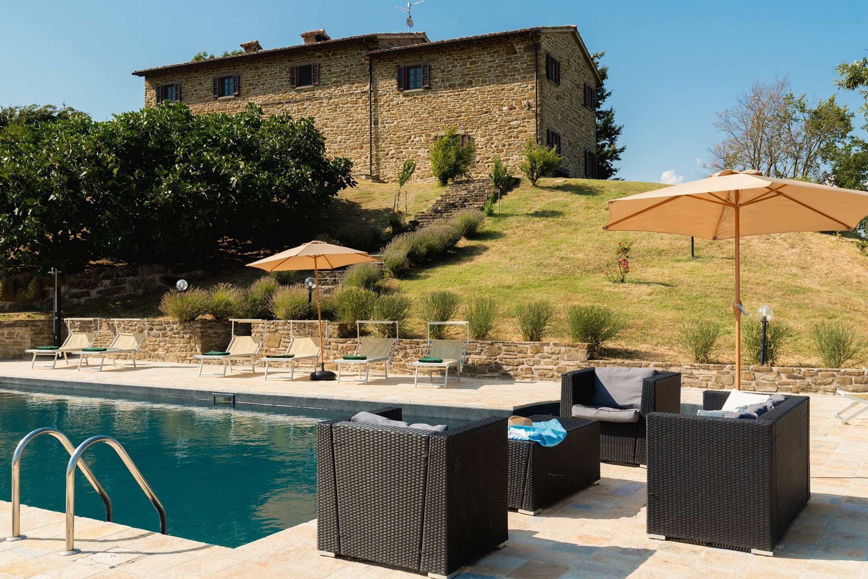 A pool area with lounge chairs and umbrellas, a stone building in the background, and a clear sky.