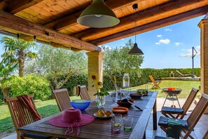 Ein Holztisch im Freien, gedeckt mit verschiedenen Gegenständen, umgeben von Stühlen, unter einer Holzpergola mit hängenden Lichtern.
