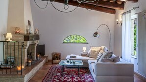 A cozy living room with a fireplace, a sofa, a coffee table, and a large window with a view. - Casale Belvedere 8 1 by Marche Holiday Villas (Pesaro)
