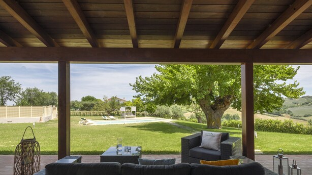 Una terrazza esterna con divano e tavolo integrati, un grande albero e vista su una zona verde con piscina sullo sfondo.