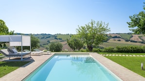 Una piscina rettangolare con superficie d'acqua cristallina e azzurra, circondata da un solarium piastrellato. Sono presenti sedie a sdraio con cuscini e un'area salotto coperta. La piscina si affaccia su dolci colline e vegetazione.