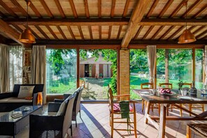 Um pátio amplo com mesa de jantar, cadeiras e sofá. Janelas grandes oferecem vista para o jardim e um edifício ao fundo.