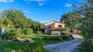 A two-story house with a tiled roof, surrounded by lush greenery and a gravel driveway. - Casale Partemio 8 in Cartoceto (Cartoceto)