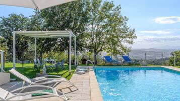 A swimming pool with lounge chairs and a pergola, surrounded by greenery and a view of distant hills.