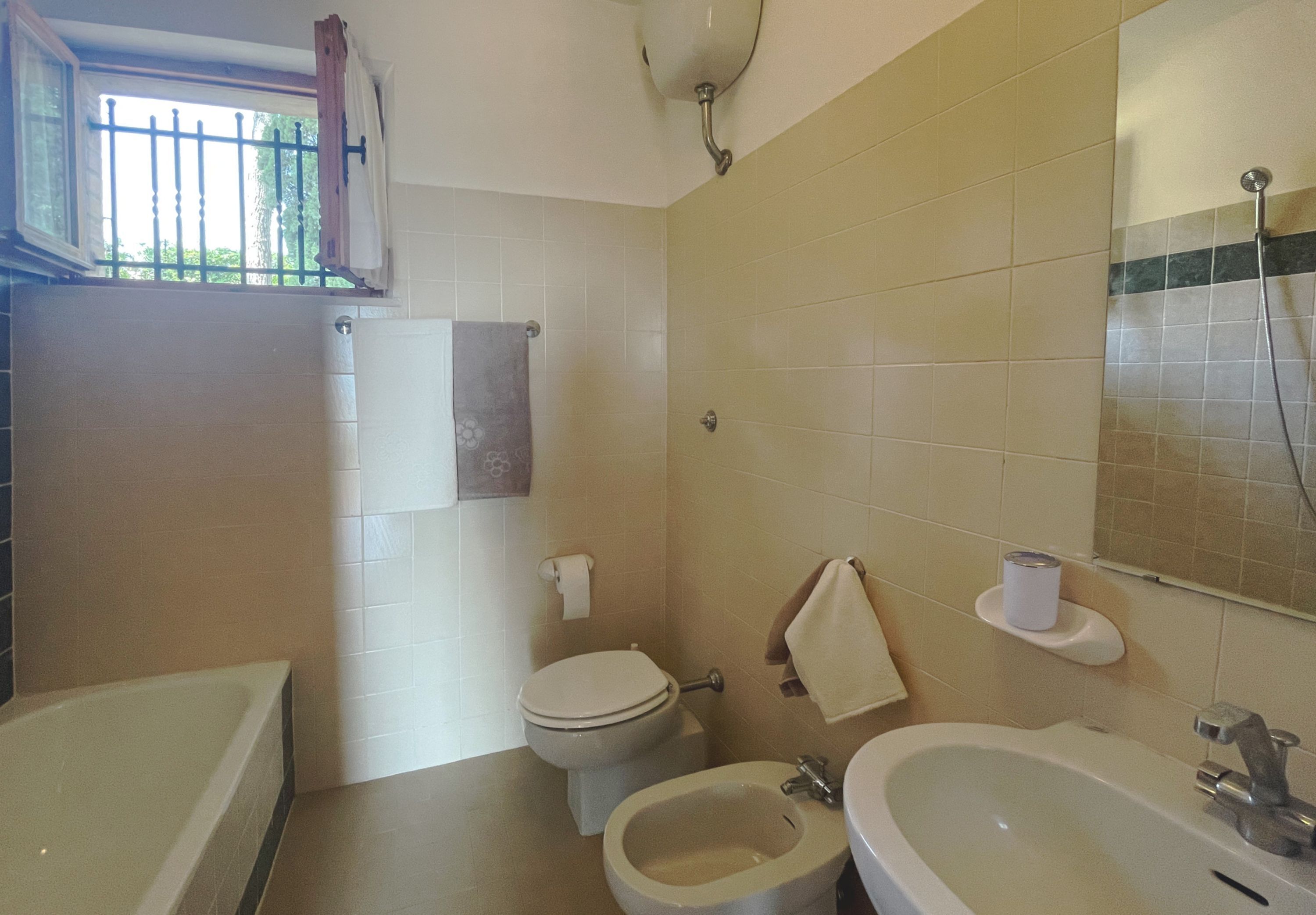 Une salle de bain équipée d’une baignoire, de toilettes, d’un bidet, d’un lavabo et d’une fenêtre donnant sur un espace verdoyant.