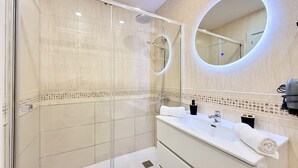 Ein modernes Badezimmer mit einer gläsernen Duschkabine, einem großen Spiegel mit runder Beleuchtung und einem weißen Waschtisch mit Handtuch.