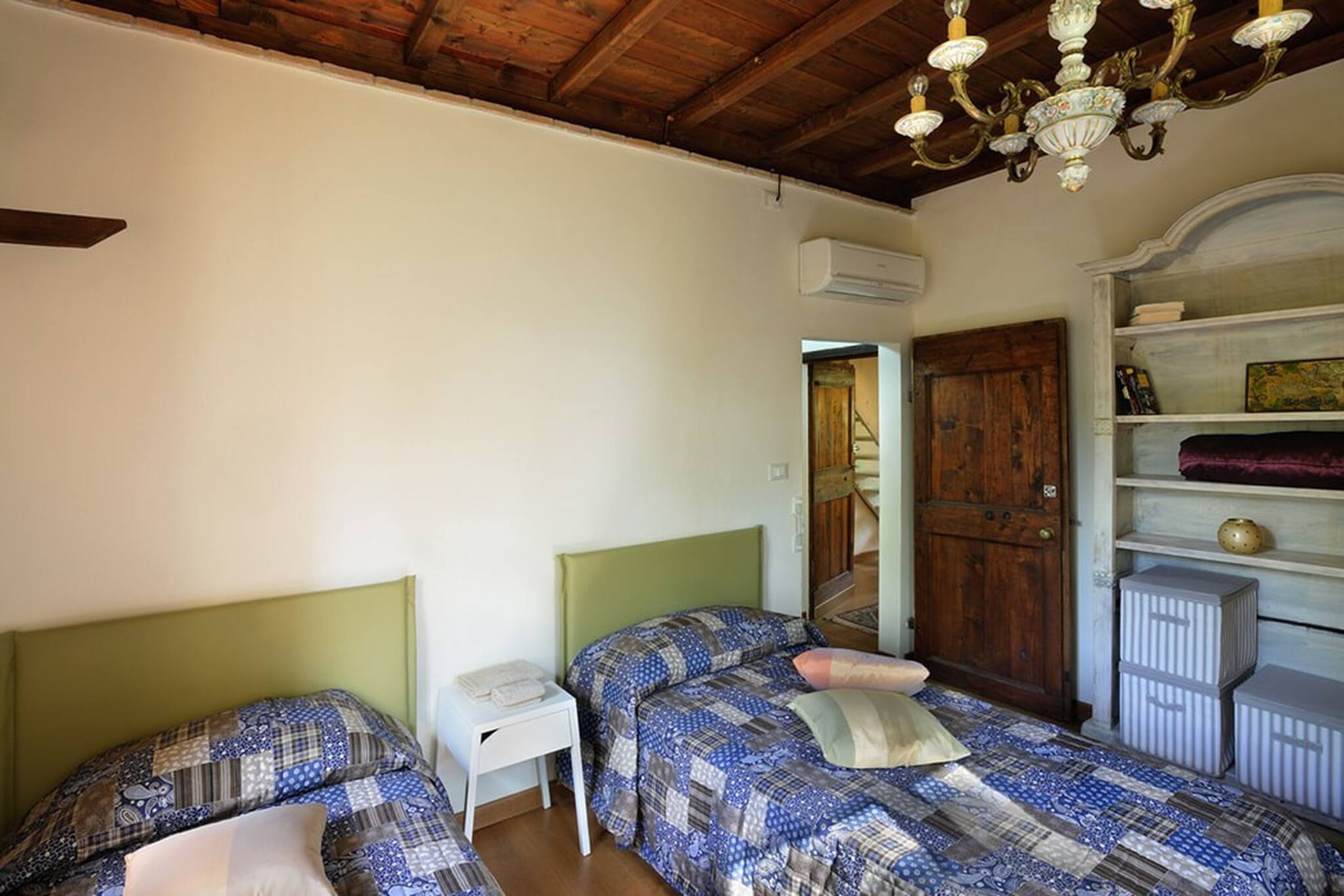A room with two beds, a wooden ceiling, a chandelier, a white wall, and a wooden door.