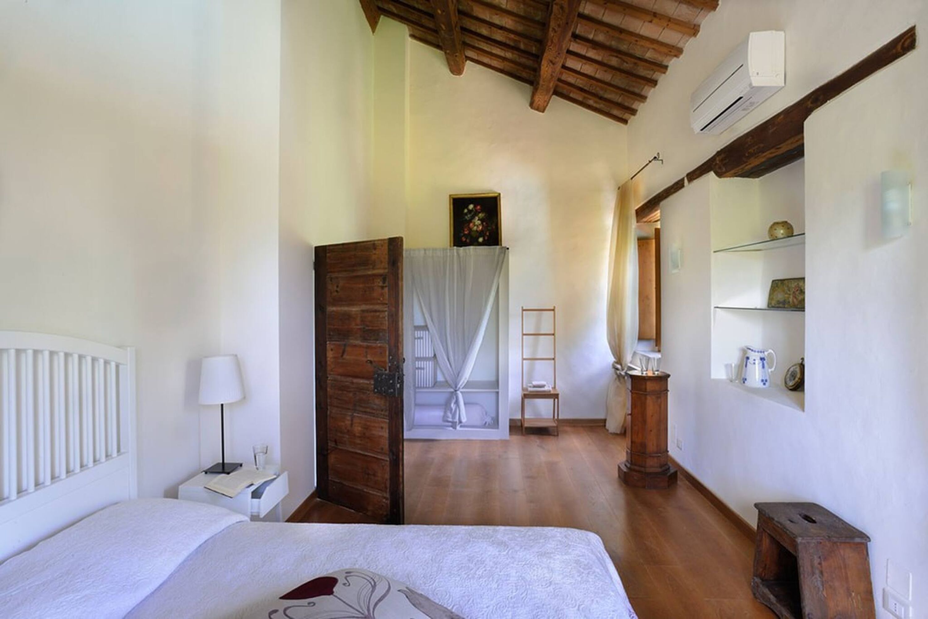 Ein Schlafzimmer mit Holzboden, einem Bett mit weißem Kopfteil, einem Nachttisch mit Lampe, einer Holztür, einem kleinen Regal mit Dekoration und einem Spiegel an der Wand.