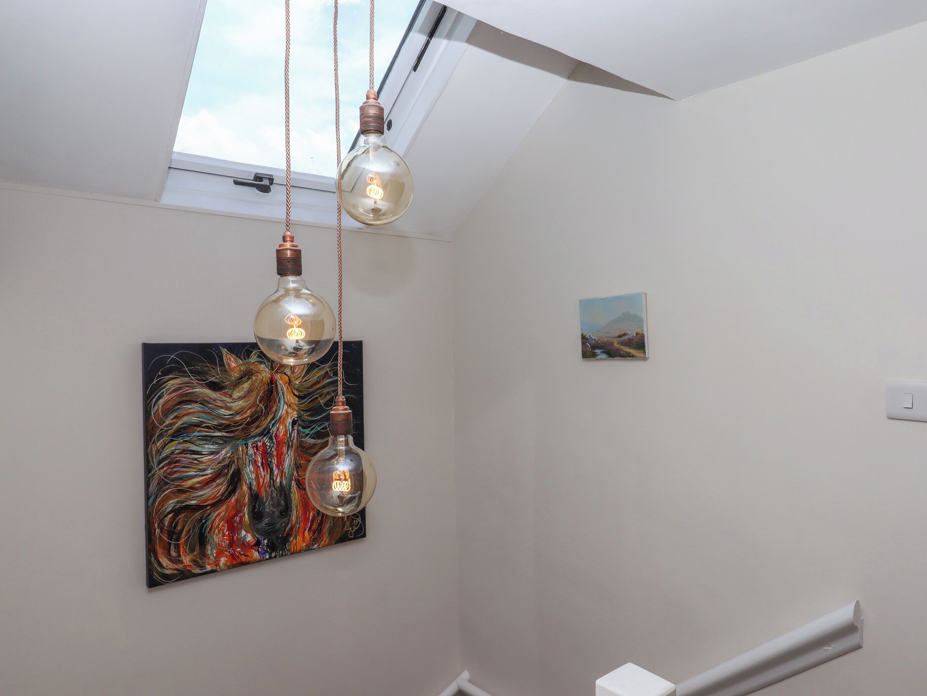 A room with a skylight, two hanging light fixtures, a colorful abstract painting, and a small landscape painting on the wall.