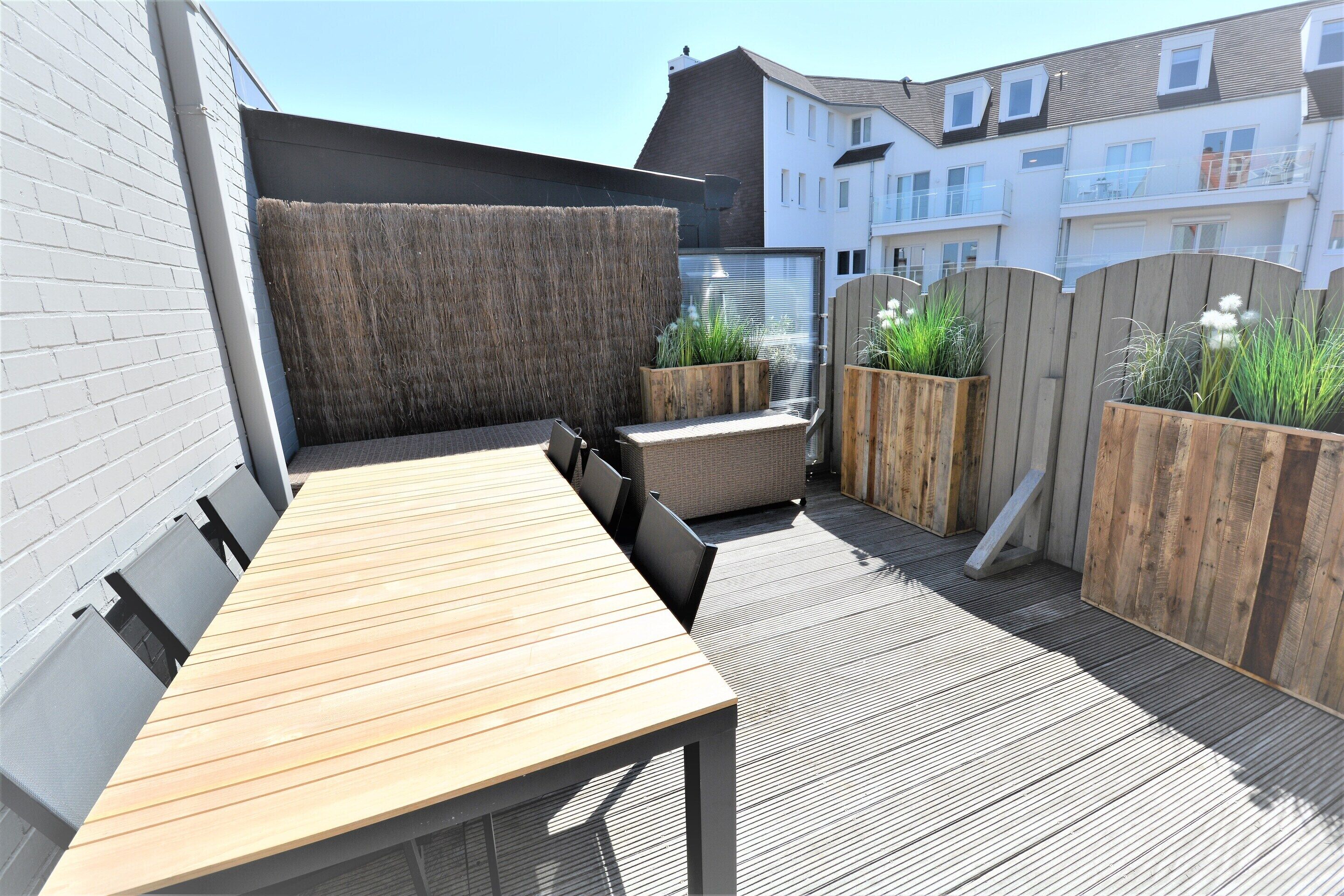 Een terras op het dak met een houten tafel en stoelen, omringd door houten plantenbakken met groen.