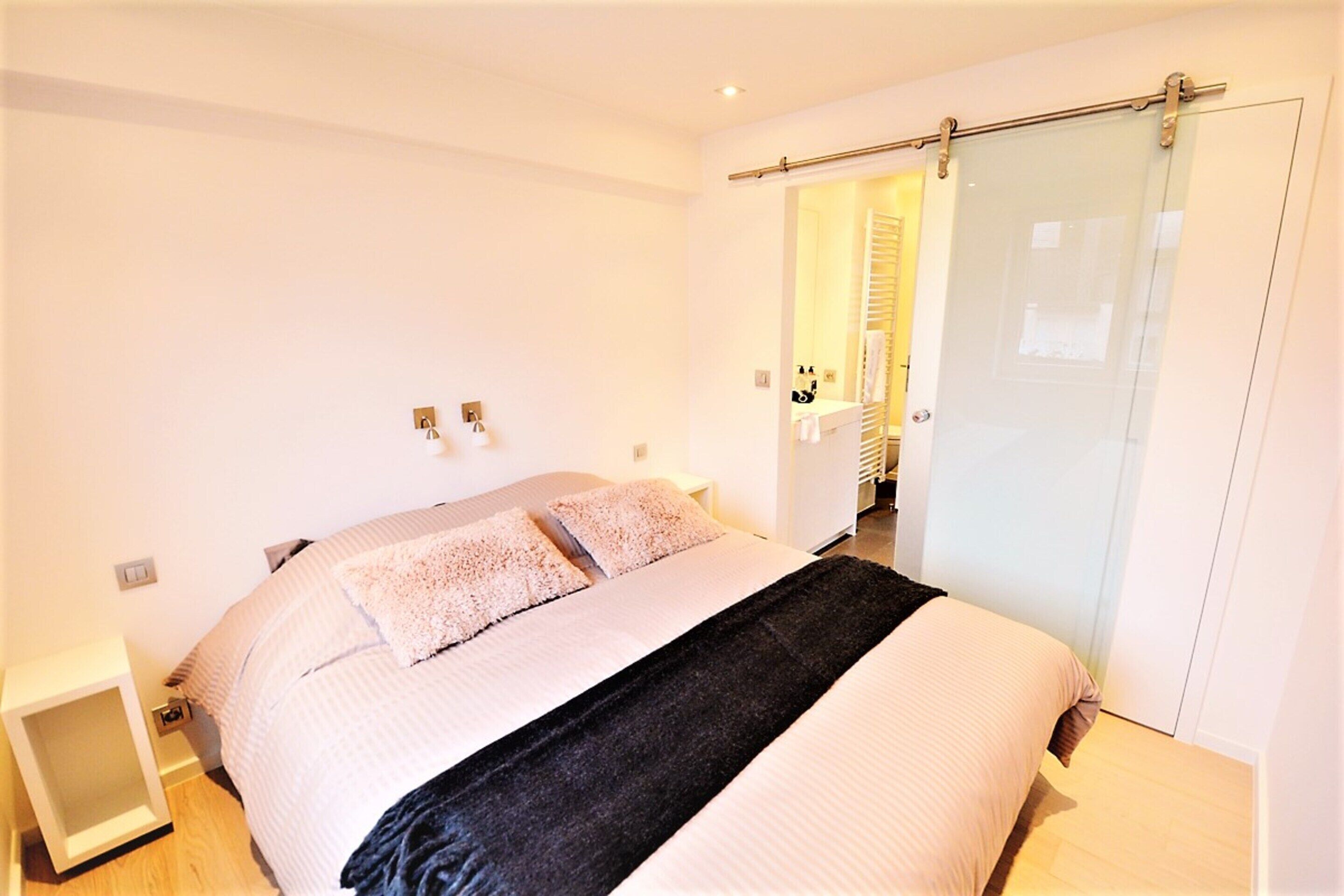 A modern bedroom with a large bed, a bedside table, and a sliding glass door leading to a bathroom.