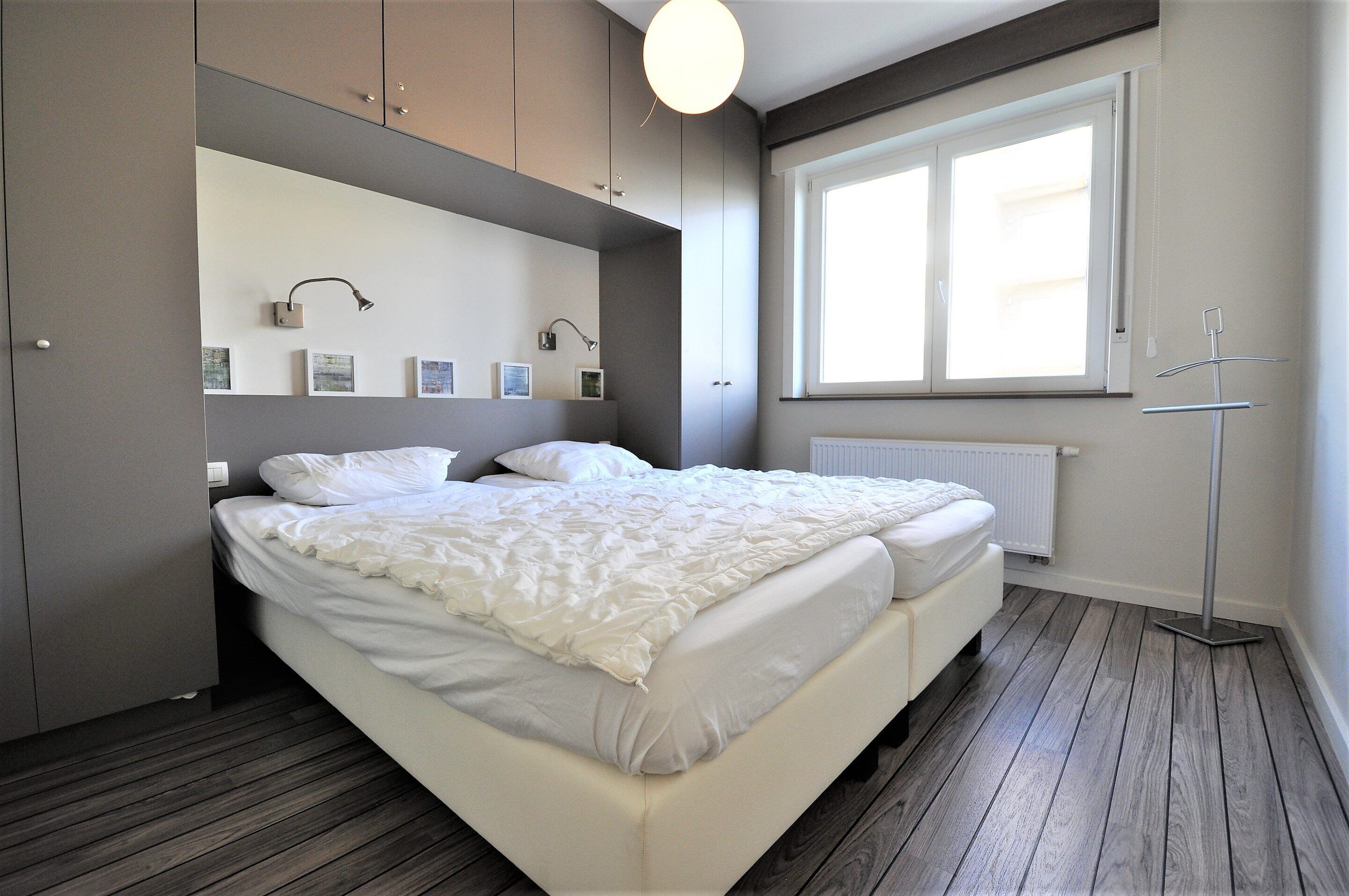 A modern bedroom with a large bed, built-in wardrobe, bedside lamps, and a window providing natural light.