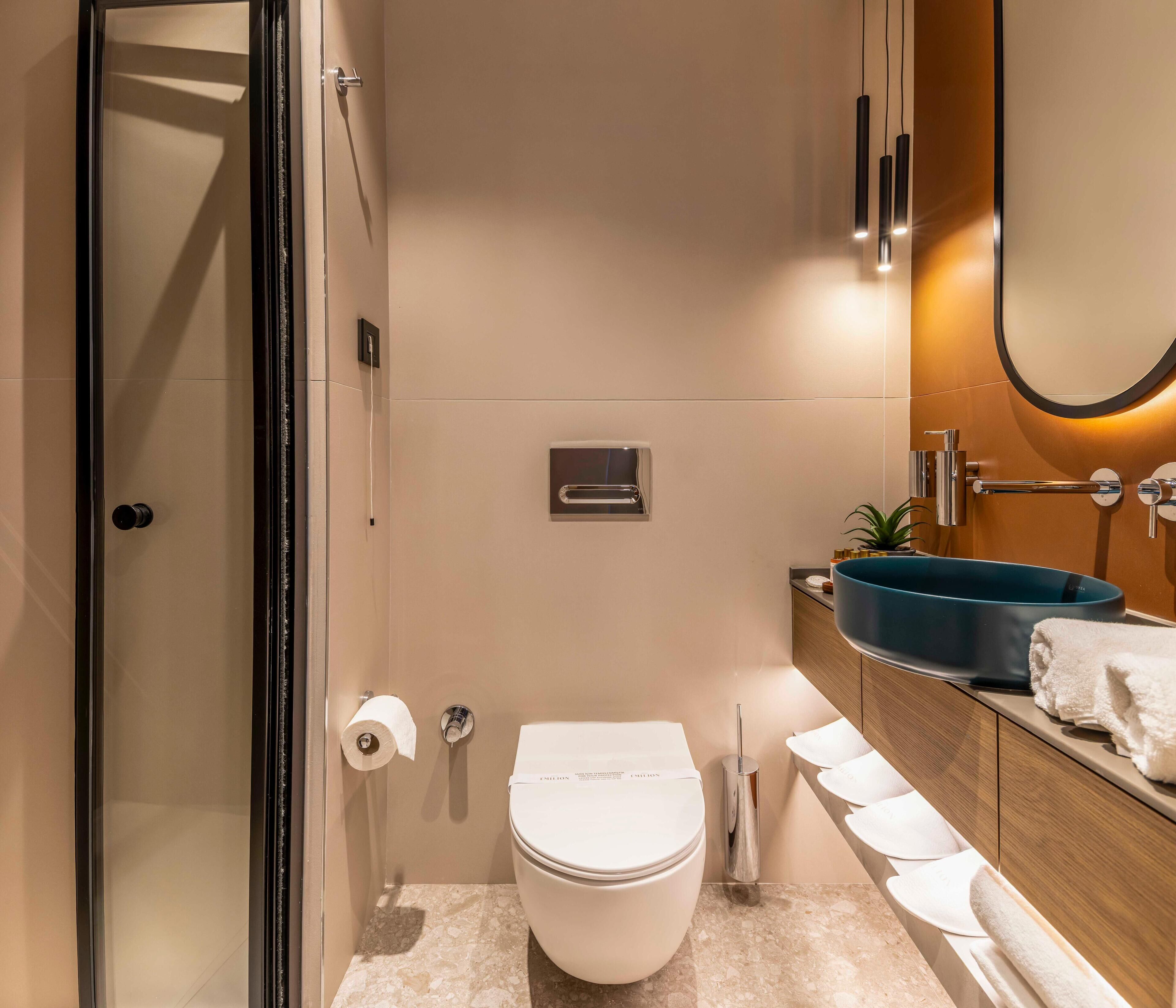 Un bagno moderno con cabina doccia in vetro, un water, un lavabo con bacino azzurro e uno specchio con cornice rotonda.