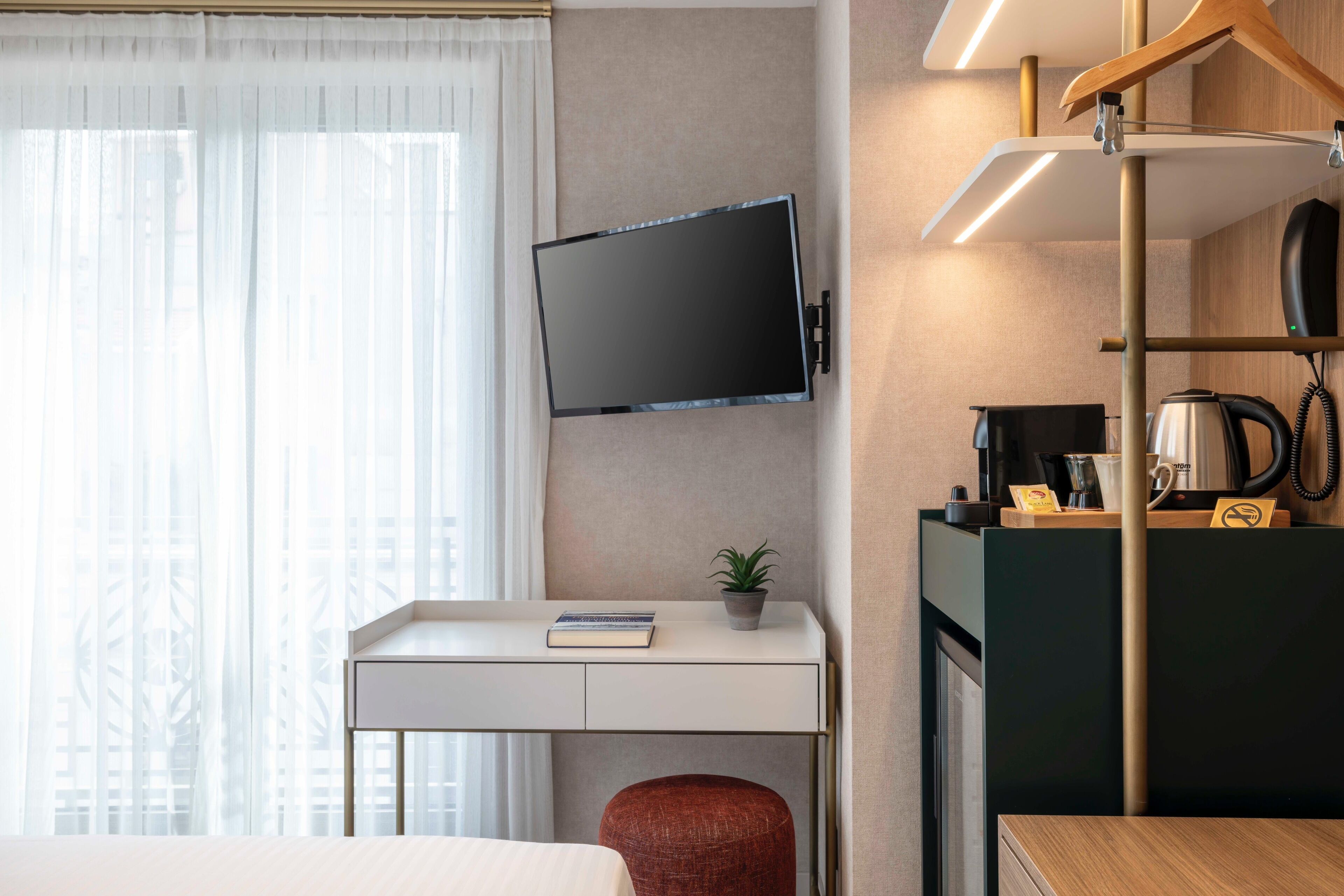 Une chambre d’hôtel équipée d’un bureau, d’un téléviseur fixé au mur, d’une petite plante, d’un pouf rouge et d’une machine à café.